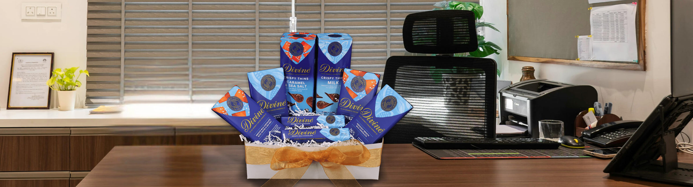 A decorative gift basket filled with blue chocolate boxes, adorned with a gold ribbon, sits on an office desk, conveying a festive and welcoming tone.