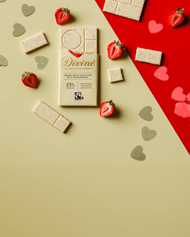 A white chocolate bar with strawberries rests on a beige and red background. Surrounded by heart shapes and strawberry halves, it evokes a romantic and sweet tone.