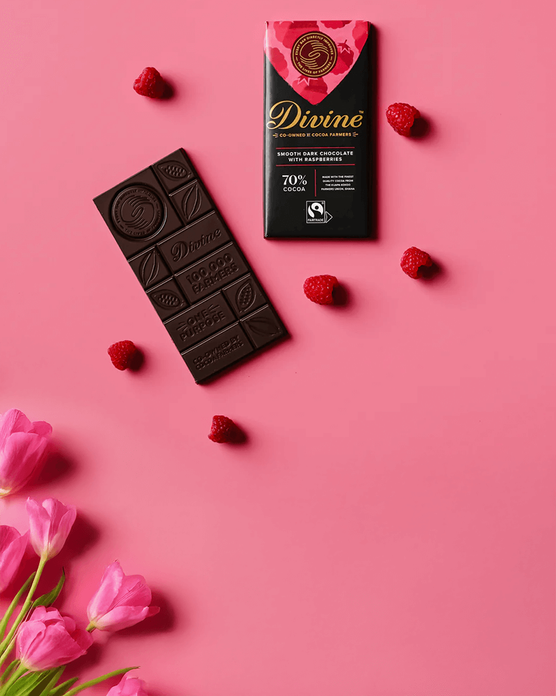 Pink background with a bar of dark chocolate next to its package, labeled "Divine" with 70% cocoa. Scattered raspberries and pink tulips create a fresh, indulgent feel.